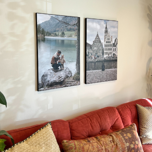 Two personalized plak-mounted prints showcasing meaningful travel moments—a lakeside snapshot and a European cityscape—tastefully installed above a red couch. A perfect blend for home decor.
