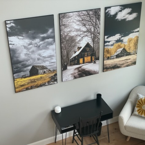 A beautiful set of three plak-mounted prints featuring rustic barn scenes through different seasons. Installed above a modern black desk setup, this wall art brings a cozy countryside charm to the workspace.
